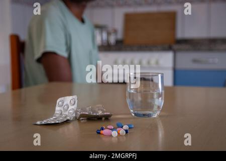 Primo piano di alcuni farmaci e un bicchiere d'acqua, persona seduta dietro, concetto di consumo di droga Foto Stock