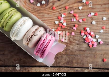 Macaron assortiti in una scatola cosparsa di piccoli cuori su uno sfondo di legno, vista dall'alto Foto Stock