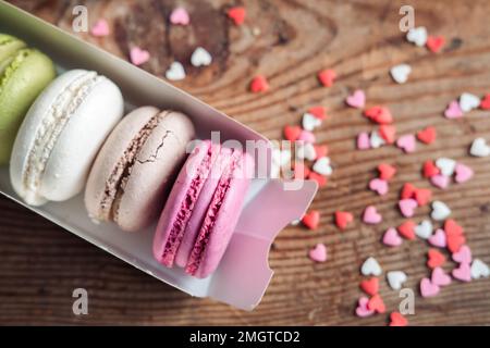 Macaron assortiti in una scatola cosparsa di piccoli cuori su uno sfondo di legno, vista dall'alto Foto Stock