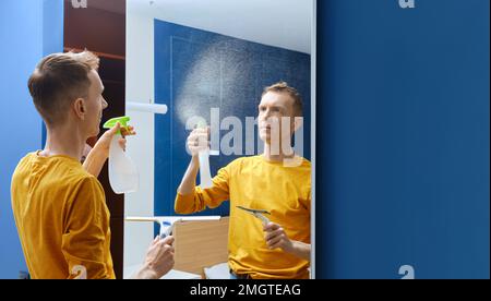 Uomo di mezza età spruzzando liquido detergente sullo specchio a casa per pulirlo Foto Stock