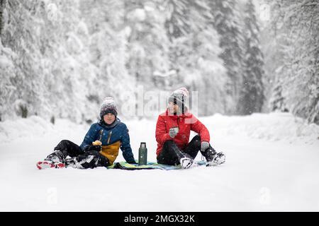 Madre e figlio, turisti, escursionisti, seduti su una coperta da picnic in una tempesta di neve pesante, divertendosi mentre bevevano tè, nelle foreste della Slovenia Foto Stock