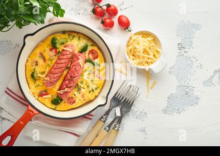 Omelette o frittata con due salsicce, formaggio fuso, pomodori ciliegini, broccoli e prezzemolo su padella rossa o teglia di ferro su fondo bianco di cemento vecchio Foto Stock