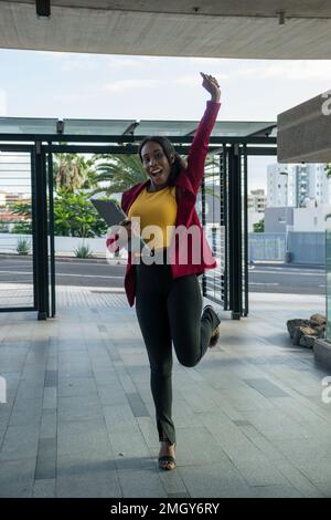 Attraente donna d'affari africana che celebra il successo aziendale tenendo il suo tablet. Immagine a tutta lunghezza Foto Stock