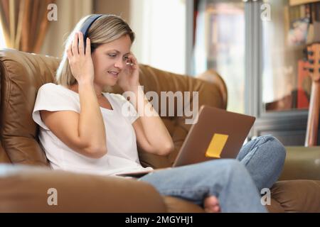 Donna sorridente con cuffie wireless e notebook seduti sul divano di casa. Foto Stock