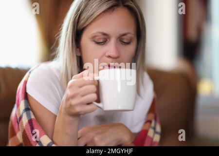 La donna gelida, avvolta in una calda coperta a scacchi, tiene a casa tazza e bevande calde. Foto Stock