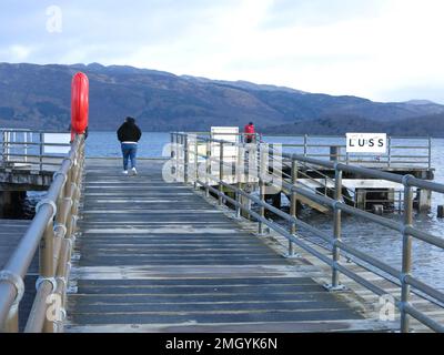 Cacciato contro il freddo, un singolo visitatore cammina lungo il molo a Luss sulle rive del Loch Lomond, in una giornata limpida nel gennaio 2023 Foto Stock