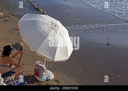 Donna che scatta foto su smartphone seduto da Parasol su Kavouri Beach Vouliagmeni Grecia Foto Stock