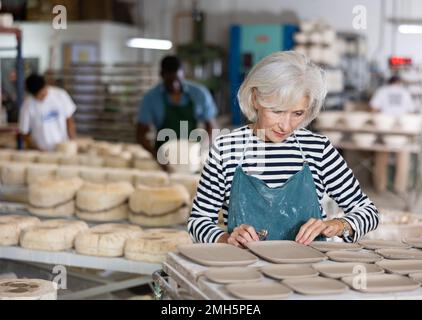 Ritratto di donna anziana in grembiule taglio bave fuori di tazze di terracotta appena fatte e piatti con coltello Foto Stock