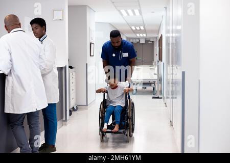 Verticale del medico afro-americano maschio che spinge il paziente del ragazzo in sedia a rotelle nel corridoio Foto Stock