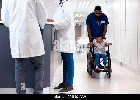 Medico afro-americano maschio che spinge il paziente del ragazzo in sedia a rotelle nel corridoio occupato dell'ospedale Foto Stock