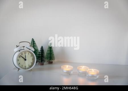 Una sveglia bianca, un puntatore a dodici, un piccolo albero di Natale artificiale verde e candele accese in candelabri su una cassettiera bianca. Copia Foto Stock