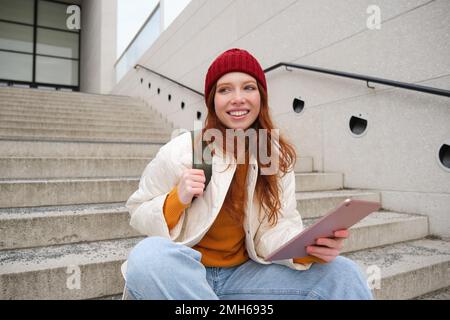 Giovane bella ragazza con lunghi capelli rossi, sorride, utilizza l'applicazione Internet, connettersi al wi-fi vicino al campus, siede sulle scale con tablet digitale Foto Stock