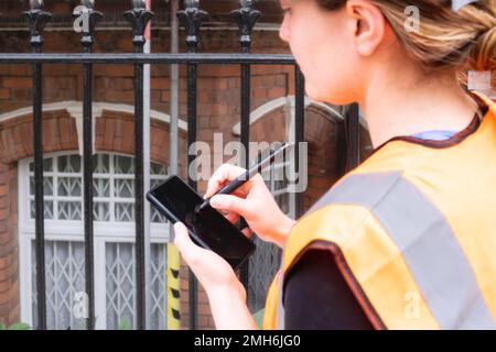 Foto ravvicinata con particolare attenzione al telefono cellulare con un ingegnere civile che prende appunti con una penna elettronica, cantiere, elmetto e ppe arancione Foto Stock