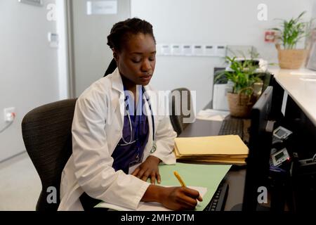 Dottore afro-americano che lavora in ospedale, seduto alla scrivania e prendendo appunti Foto Stock