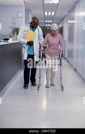Medico afro-americano maschio che cammina attraverso corridoio con paziente caucasico anziano all'ospedale Foto Stock