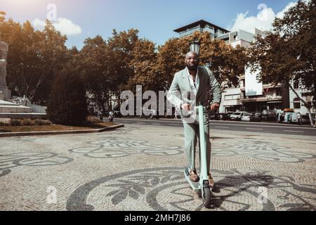 Vista di una raffinata barba baldana matura nero in sella a uno scooter elettrico sopra la pietra di pavimentazione; un elegante uomo d'affari africano in un abito sartoriale verde è ridi Foto Stock