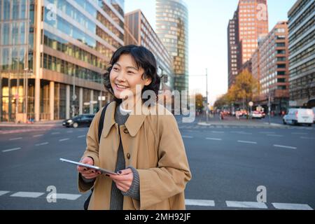 Il ritratto di una ragazza coreana sorridente si trova sulla strada trafficata del centro città, contiene un tablet digitale, un'espressione informale e rilassata del volto, camminando per la città Foto Stock