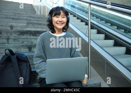 Una studentessa lavora su un progetto in remoto, si siede sulle scale, lavora su un computer portatile, ascolta la musica con le cuffie e sorride Foto Stock