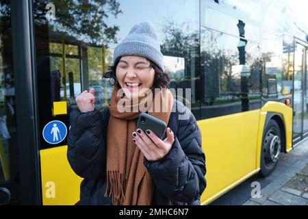 Persone moderne e stile di vita. Felice ragazza asiatica urla da gioia, festeggia, si trova vicino ai trasporti pubblici degli autobus e sembra stupito Foto Stock