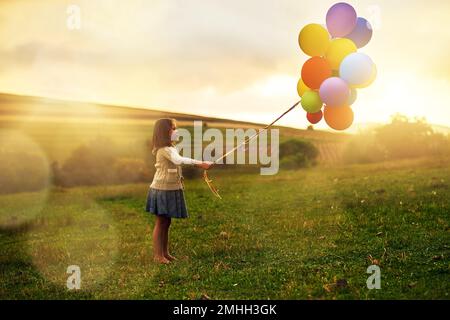 Guarda il lato luminoso. una bambina giocosa che cammina attraverso un campo mentre tiene un mazzo di palloncini. Foto Stock