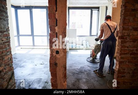 Vista posteriore di un lavoratore maschio che utilizza una macchina da trogelatura durante la spianatura del pavimento in appartamento in fase di ristrutturazione. Uomo finendo la superficie di cemento con macinacaffè per pavimenti in camera con grandi finestre. Foto Stock