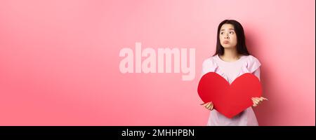 Concetto di San Valentino. Solitario adolescente asiatica ragazza sognando di amore, sentendosi triste e solitario in giornata degli amanti, guardando da parte con pietà, tenendo grande rosso Foto Stock