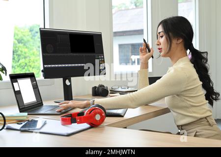 Attraente creatrice di contenuti femminile che utilizza un computer portatile e lavora con filmati e audio in uno studio creativo per ufficio Foto Stock