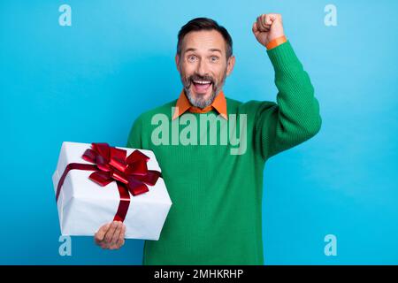 Ritratto fotografico di uomo maturo bello sollevare pugno eccitato tenere compleanno giftbox indossare abiti verdi alla moda isolato su sfondo di colore blu Foto Stock