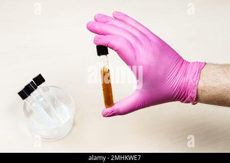 la mano con guanti può contenere la provetta con liquido marrone su un tavolo luminoso con provette con soluzioni Foto Stock