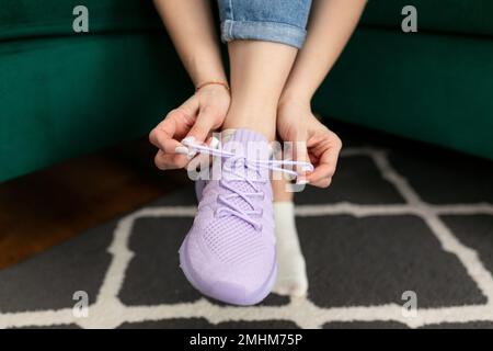 Le mani femminili pizzo su viola sneaker running seduta sul divano a casa armadio stanza. Allacciatura dei pattini, arco di legatura. Foto Stock