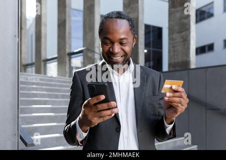 Felice e riuscito uomo afroamericano nel vestito di affari all'esterno dell'edificio dell'ufficio che tiene la carta di credito della banca e il telefono, l'uomo d'affari che acquista in linea e che trasferisce i soldi usando l'app. Foto Stock