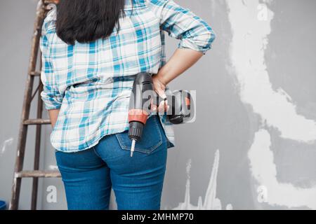 Ripresa ritagliata della schiena di una donna che tiene il trapano sui fianchi mentre guarda il muro Foto Stock