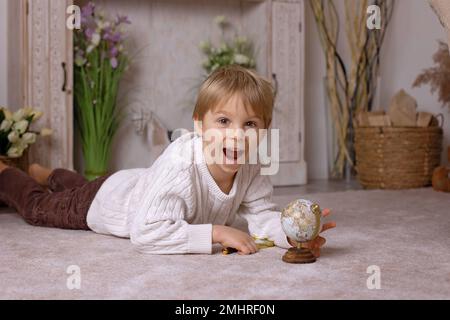 Carino bambino piccolo biondo, ragazzo, esplorando il mondo globo con il vetro magnigying, imparando i continenti Foto Stock