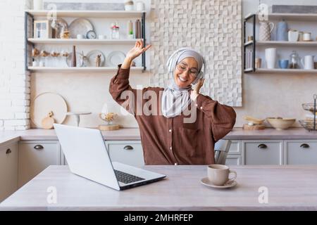 Giovane donna araba d'affari, hijabi freelancer che lavora da casa on-line a casa utilizzando il laptop. Si siede con le cuffie, ascolta la musica, alza le mani, balla, riposa, fa una pausa. Foto Stock