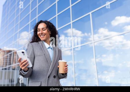 Ritratto di una giovane bella donna d'affari latino-americana in un vestito che cammina fuori vicino a un centro di vetro ufficio. Contiene una tazza di caffè, guarda il telefono, sorride. Foto Stock