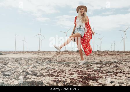 Giovane ragazza bionda che posa in un campo asciutto con alcune turbine eoliche sullo sfondo. Foto Stock