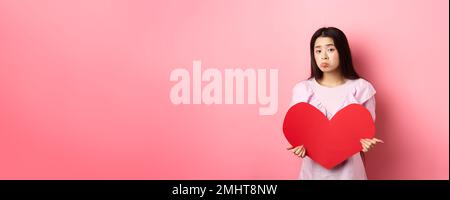 Concetto di San Valentino. Single teenage asian girl vuole innamorarsi, guardando triste e solitario alla macchina fotografica, sulking afflitto in festa degli amanti, tenendo grande Foto Stock