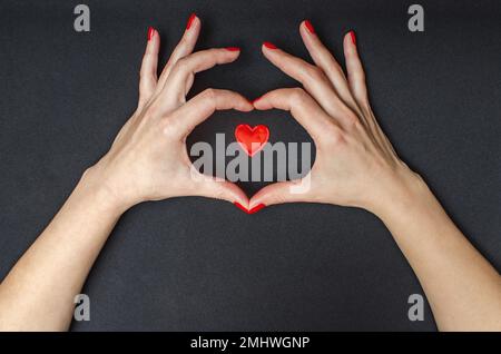 Le mani femminili intorno al cuore rosso. Amore. Sfondo nero. Smalto per unghie rosso Foto Stock