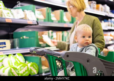 Casualy vestito madre scelta prodotti nel reparto del supermercato negozio di alimentari con il suo bambino bambino bambino bambino nel carrello della spesa Foto Stock