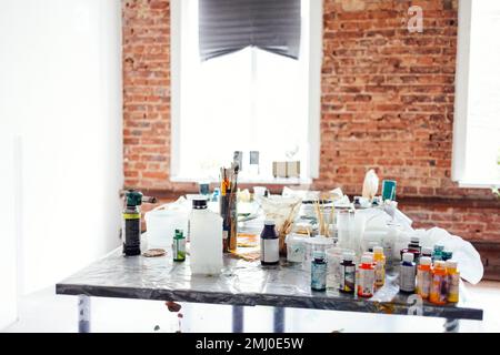 Interni moderni del laboratorio d'arte o studio. Secchielli con vernice, vasi di vetro sporchi, spazzole, pistola a spruzzo, solventi e altre forniture d'arte sono sul ar Foto Stock