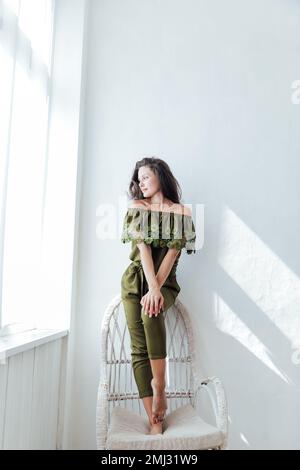 una donna in tuta verde siede accanto alla finestra su una sedia bianca Foto Stock