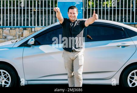 Conducente appoggiato sulla vettura mostrando la chiave, conducente maschio in piedi accanto alla vettura mostrando le chiavi con il pollice verso l'alto. Sorridente proprietario con nuova auto in piedi Foto Stock