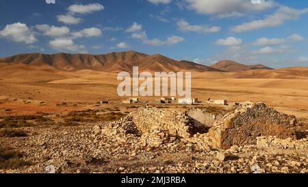 Interno dell'isola, paesaggio arido, colline brune rossastre, rovine di edifici più lontani, rovine in primo piano, cielo blu, nuvole bianche, Fuerteventura Foto Stock