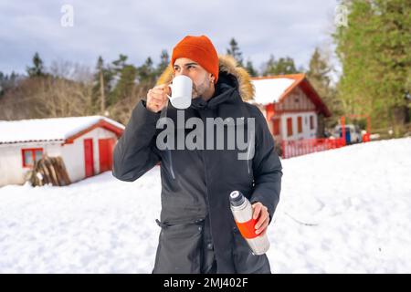 Uomo che beve il caffè da un thermos caldo in inverno nella neve accanto a una cabina Foto Stock