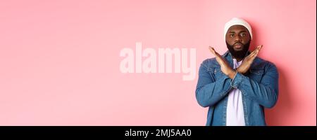Un uomo afro-americano serio che dice di no, mostrando croce per fermare qualcosa di cattivo, proibisci l'azione, in piedi su sfondo rosa Foto Stock