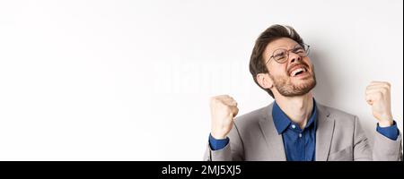 Uomo d'affari eccitato in occhiali e vestito gridando sì con piacere e volto sollevato, scuotendo pugni in su, trionfando, scommessa vincente, in piedi su bianco Foto Stock