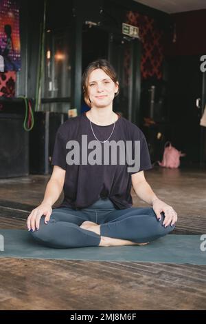 Foto verticale di una donna sorridente nuova insegnante di yoga che riposa in posa facile (sukhasana) durante la pratica di yoga con flusso di vinyasa indossando abbigliamento sportivo Foto Stock