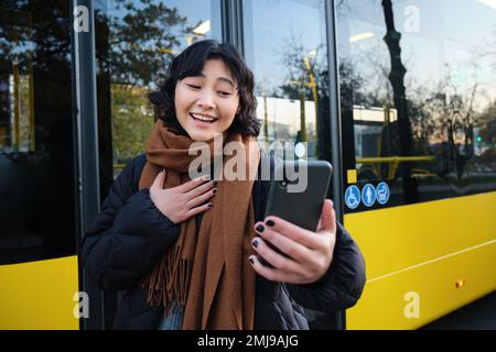 Ritratto di allegra ragazza asiatica parla sul telefono cellulare, video chat, guarda stupito smartphone fotocamera, si trova sulla fermata dell'autobus Foto Stock