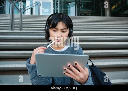 Ritratto di ragazza asiatica con il volto turbato, disegna su tablet digitale con penna, guarda complicato il suo lavoro, si sente bloccato non ispirazione, si siede sulle scale Foto Stock