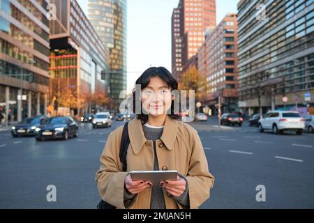 Il ritratto di una ragazza coreana sorridente si trova sulla strada trafficata del centro città, contiene un tablet digitale, un'espressione informale e rilassata del volto, camminando per la città Foto Stock